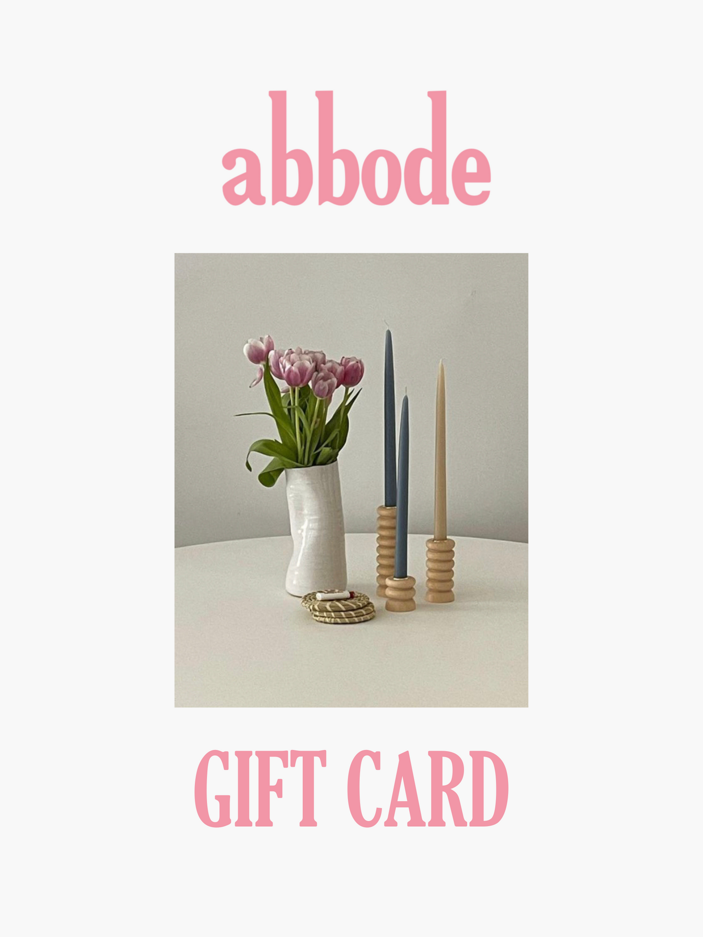 abbode gift card written in pink text. Vase filled with purple flowers sits on a table next to candles and coasters.